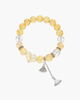 Close-up of a golden yellow citrine gemstone bead bracelet featuring a carved white heart stone and two dangling silver crystal fan charms on a mirrored surface.